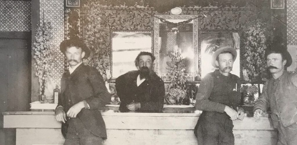 Inside the Davis Saloon at Manchester, c. 1889 — four miners at the bar under papered walls and mirrors, in a town that had a hotel, two stores, a school, and 'of course saloons.'