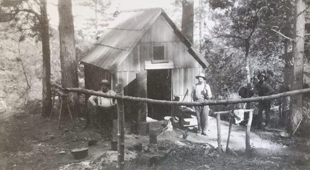 Henry Melville's mining company office at Los Burros, c. 1889 — a board-and-batten cabin in the pines, men at the rail, a dog on the step.