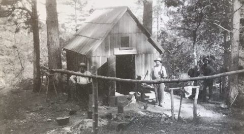 Henry Melville's mining company office at Los Burros, c. 1889 — a board-and-batten cabin in the pines, men at the rail, a dog on the step.