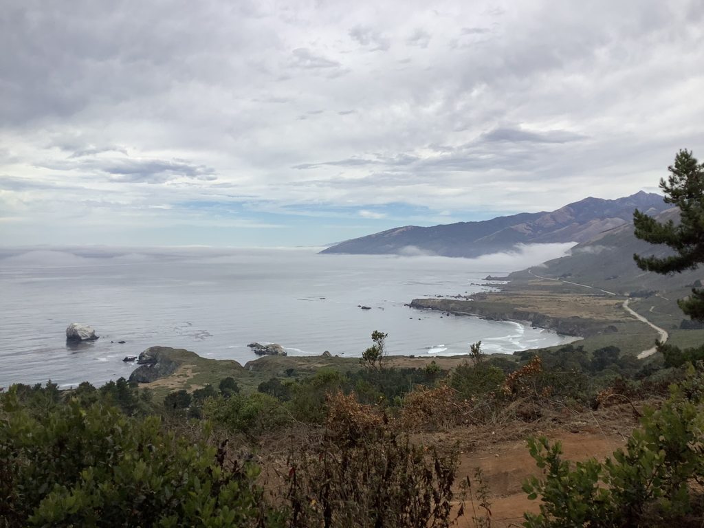 The Pacific Valley shelf from Plaskett Ridge on a grey day — Sand Dollar Beach curving below, Plaskett Rock offshore, and Highway 1 threading the family's old valley.