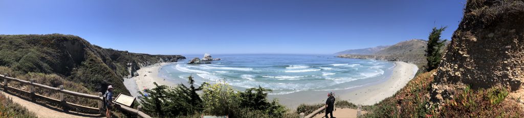 From the Sand Dollar overlook stairs: the whole crescent at once, surf stacked in rows, Plaskett Rock at the north end.