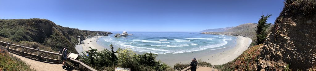 Sand Dollar panorama from the access trail — beach umbrellas below where supply boats once landed for the valley families.