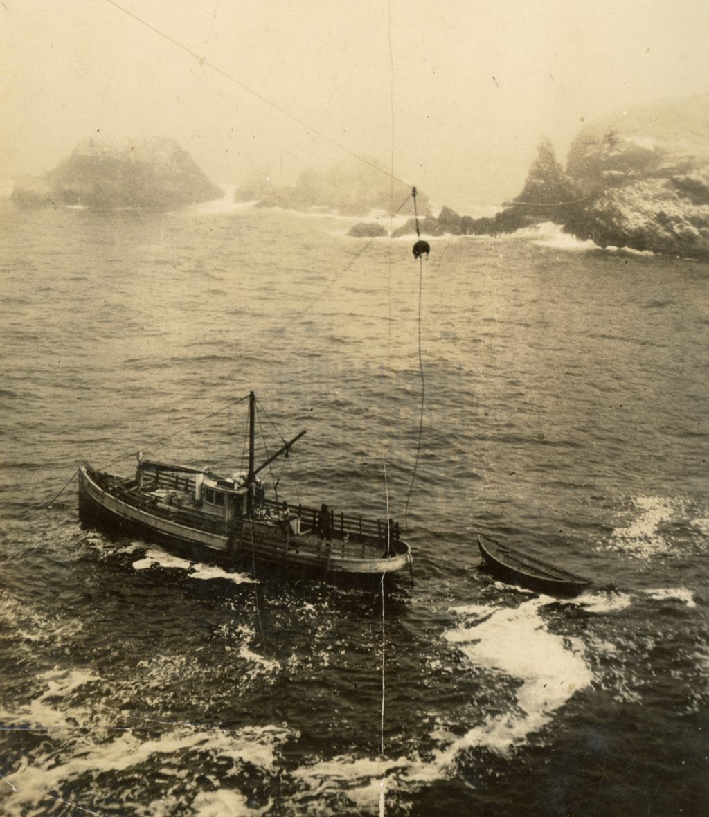 The original photograph, from the family collection: Boat Day at the Plaskett landing. The supply schooner stands off the rocks in the fog with the surf boat alongside, and on the wire between ship and bluff a hog rides the cable trolley over the water. This is the trade that supplied the South Coast — the holiday Mabel Plaskett said outshone Christmas.